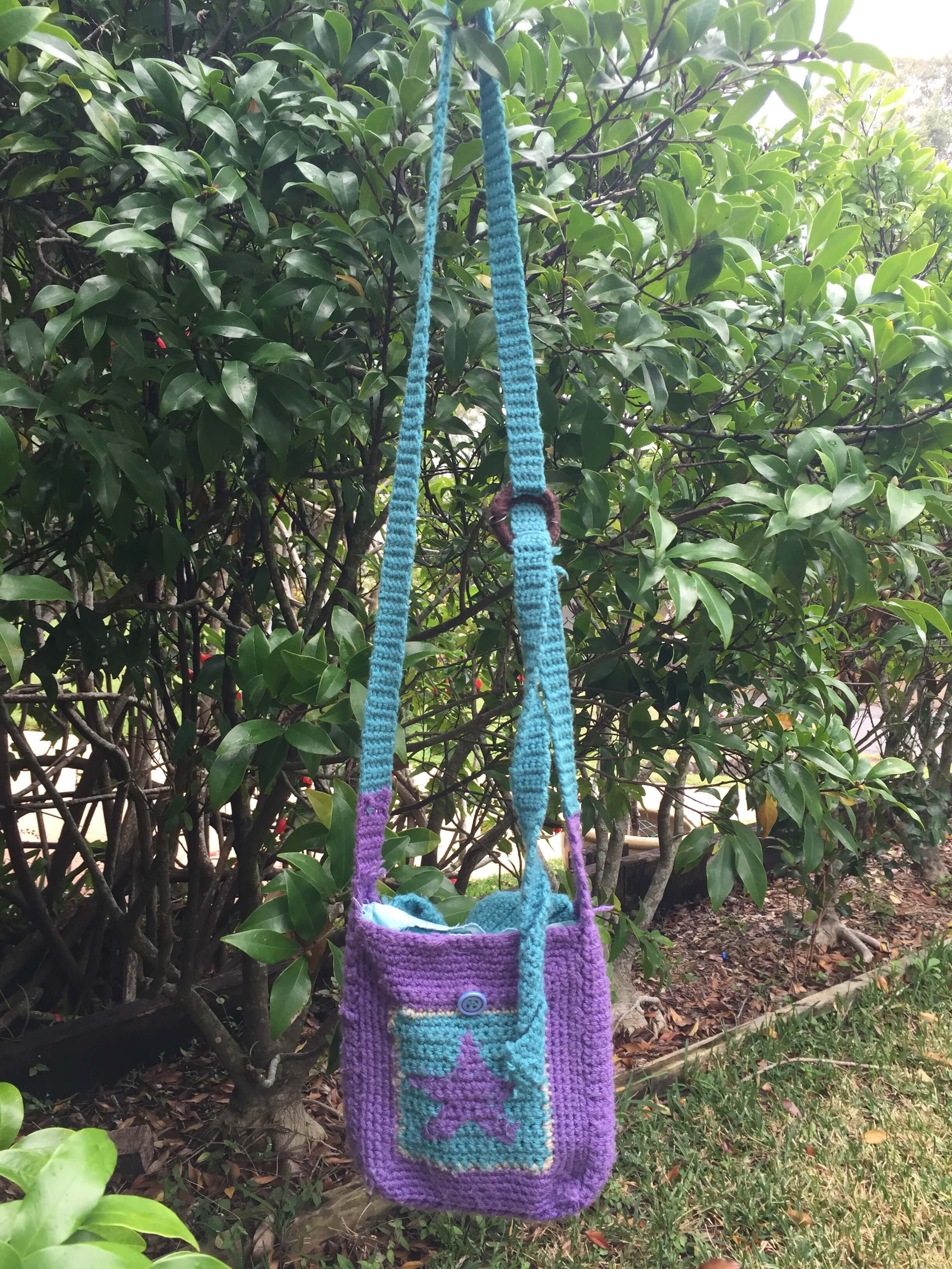 A purple and blue crocheted cross-body bag. The bag has a purple star motif on the front and a crocheted blue strap.
