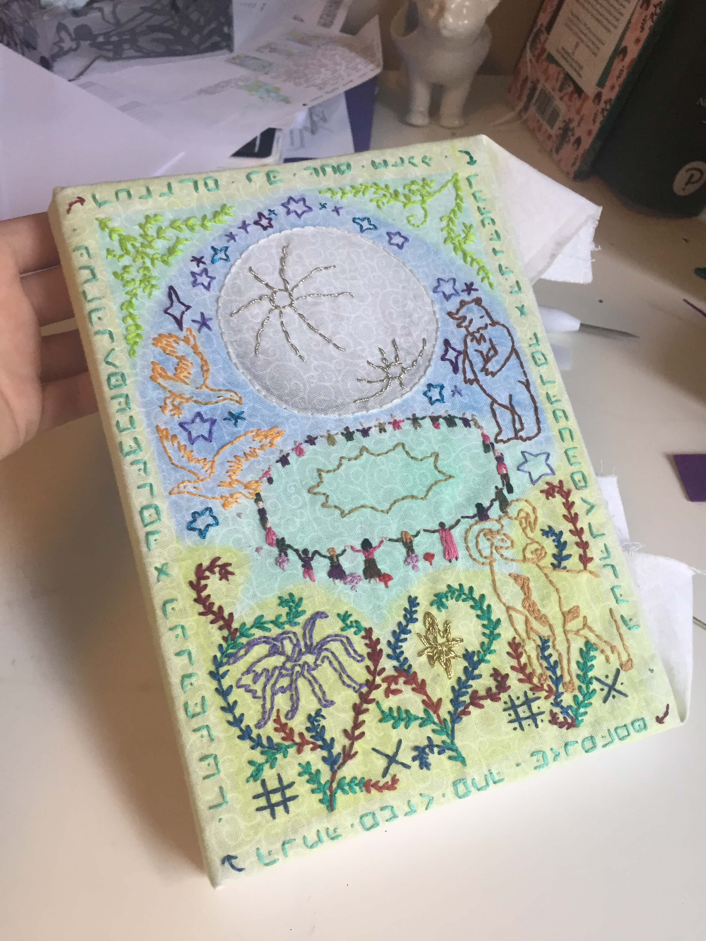 An embroidered book cover inspired by medieval illustrations. A large full moon shines in the top centre, and below it a circle of dancers revel. Embroidered ivy in colours of red, green and navy fill the corners, winding around a spider and a ram on the ground. Around the moon, stars surround an eagle and a bear. An encoded message is written around the border of the book, however I messed it up so unfortunately it can't be correctly decoded.