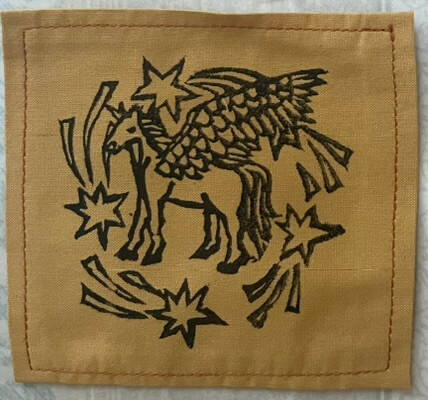 A roughly-carved black lino print on a small fabric ochre-yellow patch. The print is of a unicorn circled by shooting stars